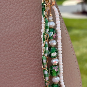 May include: A decorative bag charm featuring a gold-tone metal ring attached to a dusty pink textured surface. The charm consists of a green and white gingham ribbon, a gold chain, white seed beads, and green glass beads with floral patterns.