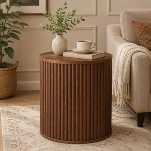 May include: A cylindrical side table crafted from dark brown wood, featuring a vertical fluted or slatted design around its exterior. The top surface is flat and circular, providing a stable base for a small ceramic vase, a book, and a cup.