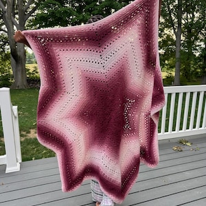 May include: A large, star-shaped crochet blanket featuring a gradient color pattern that transitions from a deep, dark burgundy at the center to lighter shades of pink and white toward the outer points, crafted with a textured, open-stitch design.