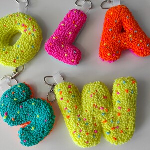 Peut inclure: Cinq porte-clés en forme de lettres tuftées O, L, A, S et W. Chaque lettre présente une texture en fil néon vibrant avec des taches multicolores ressemblant à des vermicelles. Chaque porte-clé inclut une boucle en ruban blanc et un mousqueton en métal argenté.