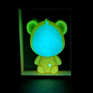 May include: A glowing bear-shaped figure encased in a rectangular frame. The bear features a bright, luminous cyan face and ears, contrasted by a yellow, textured body that emits a soft light. The entire piece is set against a dark background.