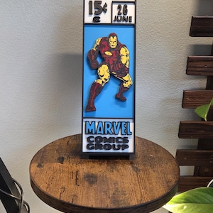 Peut inclure: Une décoration murale rectangulaire verticale présentant un design 3D en couches d'Iron Man en armure rouge et jaune sur un fond bleu clair. Le haut affiche "IRON MAN", "15¢" et "26 JUNE". Le bas indique "MARVEL COMICS GROUP".