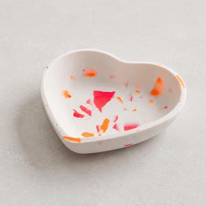 May include: A heart-shaped bowl with a white base featuring scattered, irregular flecks of bright neon orange and hot pink throughout the interior and along the outer rim, creating a speckled, multi-coloured terrazzo-style appearance.