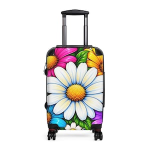 Puede incluir: Una maleta con ruedas que presenta un vibrante estampado floral con una gran margarita blanca central, rodeada de flores más pequeñas de color rosa, amarillo, azul y morado con hojas verdes. La maleta tiene un marco negro, un asa retráctil y cuatro ruedas.