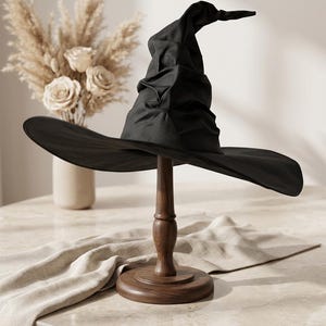 May include: A black witch hat with a pointed, slouchy crown and a wide, curved brim. The hat is displayed on a dark brown wooden stand with a circular base and a turned pedestal, showcasing the fabric texture and the structured shape of the hat.