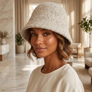 Op de afbeelding: Een lichtgrijze en beige gespikkelde vilten bucket hat met een afgeronde kroon en een naar beneden lopende rand. De hoed heeft een getextureerd, wolachtig uiterlijk met een zacht, gemêleerd kleurpatroon over de gehele stof van het hoofddeksel.