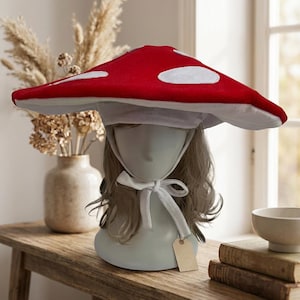Peut inclure: Un chapeau à larges bords en forme de chapeau de champignon, doté d'un dessus rouge vif avec des taches ovales blanches et un dessous blanc. Le chapeau comprend une mentonnière blanche avec des liens en tissu sur un mannequin à cheveux clairs.