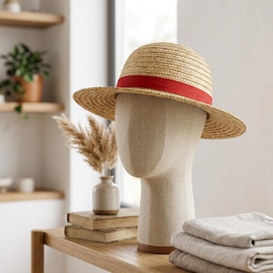 May include: A light tan straw hat with a rounded crown and a wide brim, featuring a vibrant red ribbon band encircling the base of the crown. The hat is displayed on a neutral-coloured mannequin head, showcasing the woven texture of the straw material.