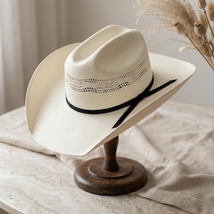 May include: A cream-colored cowboy hat featuring a woven straw texture, a cattleman crease crown, and a wide brim. The hat is accented with a thin black ribbon band tied in a small bow and a decorative perforated pattern around the base of the crown.