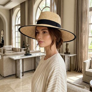Puede incluir: Un sombrero de paja de ala ancha con una textura tejida en tono beige natural, una copa estructurada y una cinta negra sólida que rodea la base. El ala está rematada con un borde negro distintivo en el contorno, creando un contraste.
