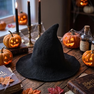 Peut inclure: Un chapeau pointu noir à large bord, doté d'une construction en tricot texturé. Le chapeau est placé au centre, entouré de citrouilles sculptées, de bougies noires allumées dans des bougeoirs en laiton et de vieux livres sur une surface en bois.