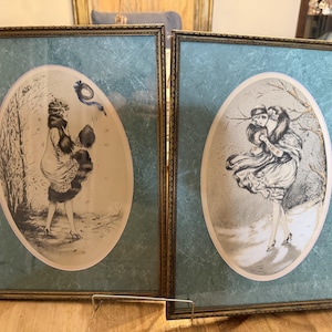 May include: Two rectangular framed prints featuring oval-shaped black and white illustrations of figures in fur-trimmed clothing. Each print is set against a textured teal background within a thin, metallic gold-toned frame with decorative edges.