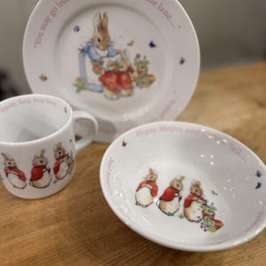 May include: A white ceramic three-piece dinnerware set featuring illustrations of rabbits in red jackets. The plate, bowl, and mug include printed text such as "You may go into the fields or down the lane" and "Flopsy, Mopsy, and Cotton-tail."