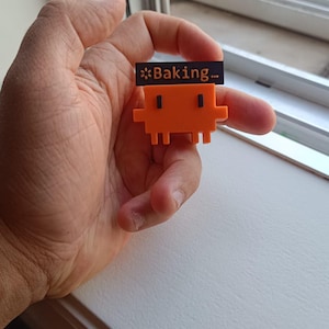 Pode incluir: Uma pequena figura de plástico laranja brilhante com um design pixelizado e quadrado. A figura apresenta dois pequenos olhos retangulares pretos e uma placa retangular preta fixada no topo com o texto Baking... em fonte branca.