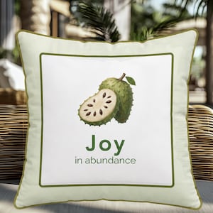 May include: A square throw pillow with a light green border and a white centre panel. The design features a green soursop fruit, with one half sliced open to reveal white flesh and black seeds. Below the fruit, the text reads Joy in abundance.