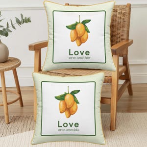 May include: Two square throw pillows with a light cream background and a thin gold border. Each pillow features a central white square containing a watercolour illustration of yellow mangoes with green leaves and the text Love one another.