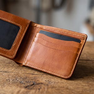 Può includere: Un portafoglio bifold in pelle color cuoio con cuciture chiare a vista, molteplici scomparti interni per carte e una finestra portadocumenti foderata in rete nera. Il portafoglio è aperto e mostra i suoi scomparti interni e una carta nera.