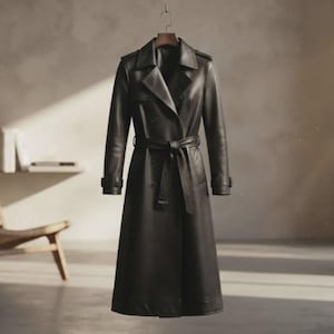 Könnte beinhalten: Ein langer Trenchcoat aus dunkelbraunem Leder mit breitem Reverskragen, langen Ärmeln mit Schnallenmanschetten und einem passenden Bindegürtel in der Taille. Der Mantel hat einen zweireihigen Verschluss und Schulterklappen und hängt an einem Holzbügel.