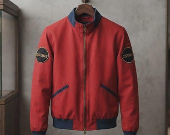 Könnte beinhalten: Eine rote Bomberjacke mit dunkelblauem Kragen, Bündchen und Saum. Die Jacke verfügt über einen durchgehenden Frontreißverschluss und zwei schräge Seitentaschen. Auf jedem Ärmel befindet sich ein runder schwarzer Aufnäher mit dem Wort CUSTOMIZE in Gold.