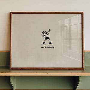 May include: A framed print with a light gray background featuring a small black line drawing of a person playing tennis and the text "he's a tennis boy" written in a simple cursive font below the figure, enclosed in a dark brown wooden frame.