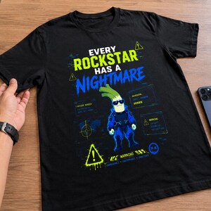 May include: A black short-sleeved t-shirt featuring a graphic print with the text "EVERY ROCKSTAR HAS A NIGHTMARE". The design includes a central character wearing sunglasses, surrounded by digital interface elements, warning icons, and blue and neon green accents.