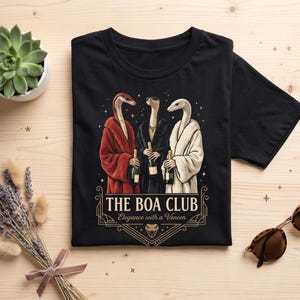Può includere: Una t-shirt nera con una grafica di tre serpenti in accappatoio che tengono bottiglie di champagne. Il testo recita THE BOA CLUB ed Elegance with a Venom, accompagnato da un'icona di testa di serpente, il tutto in una cornice geometrica dorata.