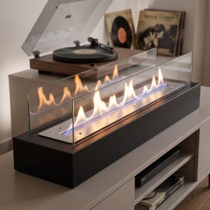 May include: A rectangular tabletop fireplace with a matte black base and a clear glass wind guard. The burner features a long, narrow metal insert with active orange and blue flames rising from the centre, creating a linear fire display.