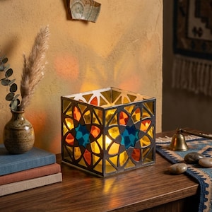 May include: A square glass lantern featuring a dark metal frame with an intricate geometric cutout pattern. The panels are filled with translucent stained glass in shades of blue, orange, and yellow, illuminated from within by a warm light source.