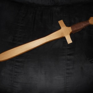 May include: A wooden toy sword with a brown leather wrap on the handle. The sword is about 24 inches long and has a rounded tip.