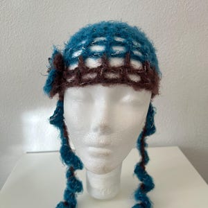 Peut inclure: Un bonnet crocheté bleu sarcelle et marron avec un motif en grille et des fils argentés métalliques. Le bonnet présente une fleur crochetée décorative sur le côté et deux longues tresses crochetées en spirale qui pendent.