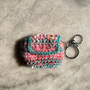 Peut inclure: Une petite pochette rectangulaire en crochet avec un motif de fil panaché dans des tons de rose, bleu sarcelle et blanc. La pochette possède un rabat et est fixée à un porte-clés en métal argenté avec un fermoir mousqueton.