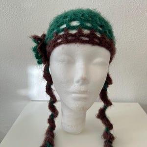 Peut inclure: Un bonnet en crochet avec une calotte verte à mailles ouvertes et une bande marron. Une fleur en crochet marron est fixée sur le côté gauche. Deux longs liens tressés en spirale marron et vert pendent sur les côtés du bonnet.