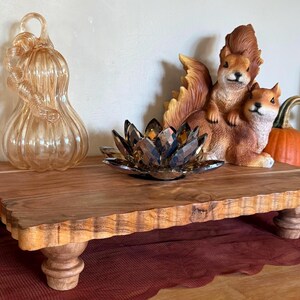 May include: A rectangular wooden riser with a scalloped edge and four turned feet. On the riser sits a dark amber crystal lotus flower sculpture, a clear glass pumpkin, and a figurine of two brown squirrels. A pumpkin is visible on each side.