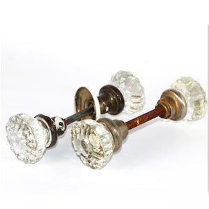 May include: Two vintage door knobs featuring clear, faceted glass handles with a starburst pattern. Each knob is attached to a dark, weathered metal base and a long, slender metal spindle. The metal components show signs of age and oxidation.