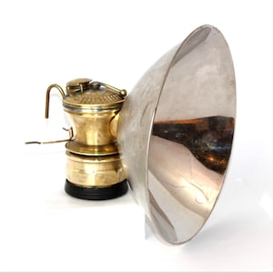 May include: An antique brass miner's lamp with a large, silver-colored reflector. The lamp has a cylindrical body with a handle and a small spout. The reflector is a large, curved metal piece. The lamp sits on a black base.