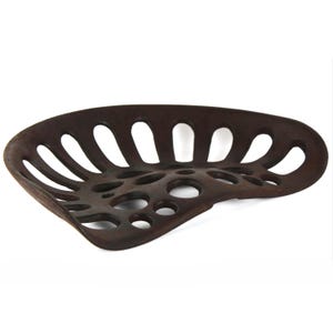 Könnte beinhalten: Ein Vintage-Traktorsitz in Braun mit geschwungenem Design. Der Sitz besteht aus Metall und weist eine Reihe ovaler und kreisförmiger Aussparungen zur Belüftung auf. Der Sitz hat ein verwittertes Aussehen, was auf Alter und Nutzung hindeutet. Der Sitz ist etwa 40 cm breit.