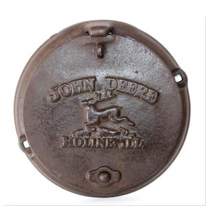 Puede incluir: Una tapa de alcantarilla redonda de hierro fundido marrón vintage John Deere con el logotipo de John Deere y el texto "Moline, Ill."
