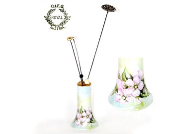 May include: A porcelain vase with a flared base, painted with light pink apple blossoms and green leaves on a pale blue and white background. The top features three thin black metal stems with small decorative gold and white accents.
