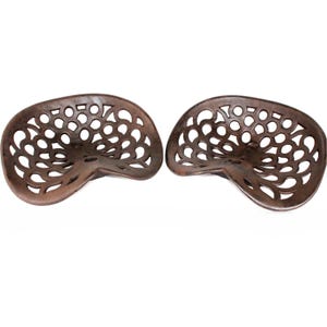 Puede incluir: Dos asientos de tractor antiguos de hierro fundido. Los asientos son marrones con un acabado envejecido y presentan un patrón decorativo de recortes circulares. Los asientos están diseñados para brindar comodidad y soporte al usuario.