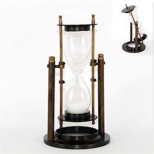 Puede incluir: Un reloj de arena giratorio con un bulbo de vidrio transparente lleno de arena blanca, sostenido por un marco de metal en tono latón oscuro. La estructura incluye una base circular, dos pilares de soporte verticales y un mecanismo pivotante.