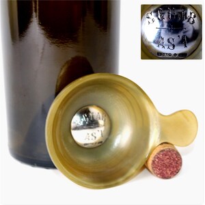 Puede incluir: Un embudo de cuerno translúcido de color claro con un asa pequeña y un inserto central de metal plateado grabado con el texto "SCUAB AS'I" y "W.D&Co". El embudo está colocado junto a una botella de vidrio oscuro y un tapón de corcho.