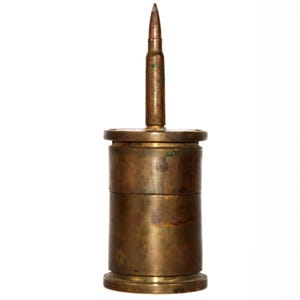 May include: A cylindrical brass container with a weathered, tarnished finish. The lid features a centered, upright rifle cartridge casing that acts as a handle. The object has a two-part construction with a visible seam around the middle.