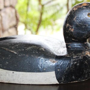 May include: A black and white wooden duck decoy with a white stripe around the middle. The decoy has a black eye with a yellow iris.