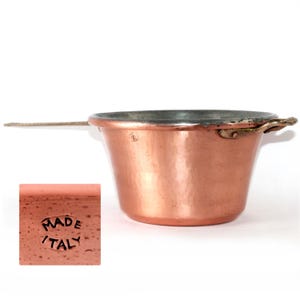 Puede incluir: Una cacerola de cobre con un mango largo y dos asas laterales pequeñas. La olla tiene una textura martillada y un borde oscuro. Un pequeño bloque de cobre con las palabras "MADE ITALY" está en la esquina inferior izquierda.