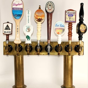May include: A brass beer tap display featuring eight different tap handles. The handles have various designs and logos, including "Blatz", "Leinenkugel's Toasted Bock", "New Glarus", "Summer Ale", "George Killian's Irish Red", "Last Lake Amber", "Gray's Oatmeal Stout", and "Genuine Draft Light".