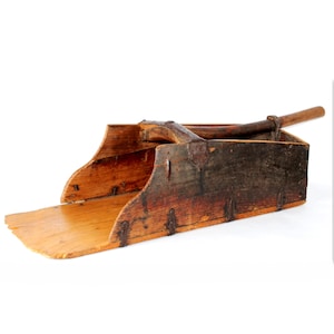 Puede incluir: Una antigua pala de grano de madera con un mango largo. La pala tiene una forma rectangular con una base ancha y plana y lados curvos. La madera está envejecida con un aspecto oscuro y desgastado, mostrando signos de uso y edad.