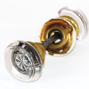 May include: A clear glass door knob set featuring a round, faceted center design. The knobs are attached to a central metal spindle and include circular brass-toned mounting plates with visible screw holes and signs of aged patina.