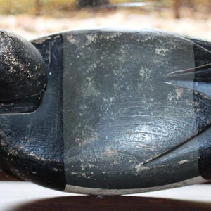 May include: A black wooden duck decoy with a detailed carving of the head and feathers. The decoy has a worn, aged look and is a great example of vintage hunting decor.