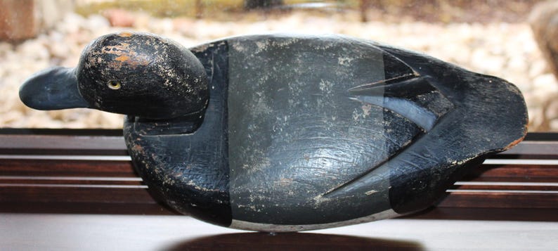 May include: A black wooden duck decoy with a detailed carving of the head and feathers. The decoy has a worn, aged look and is a great example of vintage hunting decor.