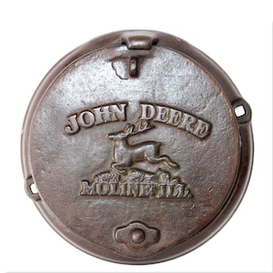 Puede incluir: Una tapa redonda de hierro fundido marrón con un logotipo de John Deere. El logotipo presenta un ciervo saltando con cuernos y las palabras "John Deere" sobre el ciervo y "Moline, Ill." debajo del ciervo.